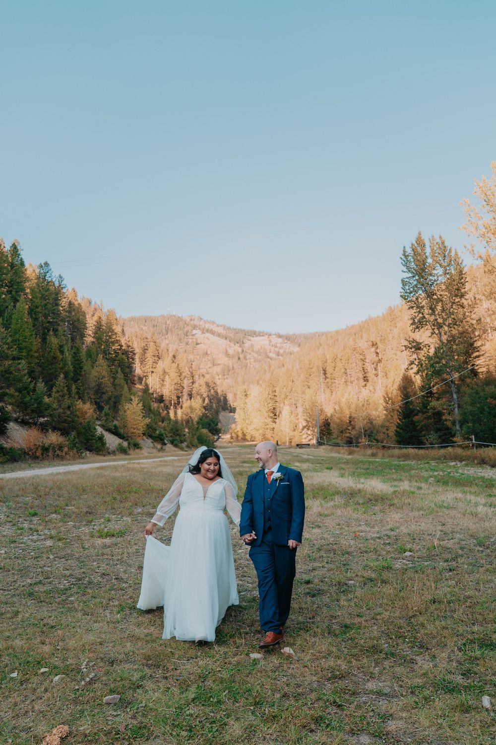 Bride and groom walk hand in hand through a sunlit mountain meadow surrounded by autumn trees on their wedding day.