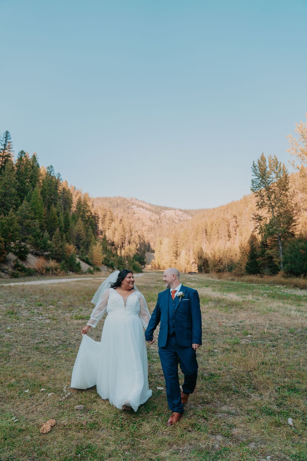 Bride and groom walk hand-in-hand through a mountain meadow surrounded by pine trees on their wedding day.