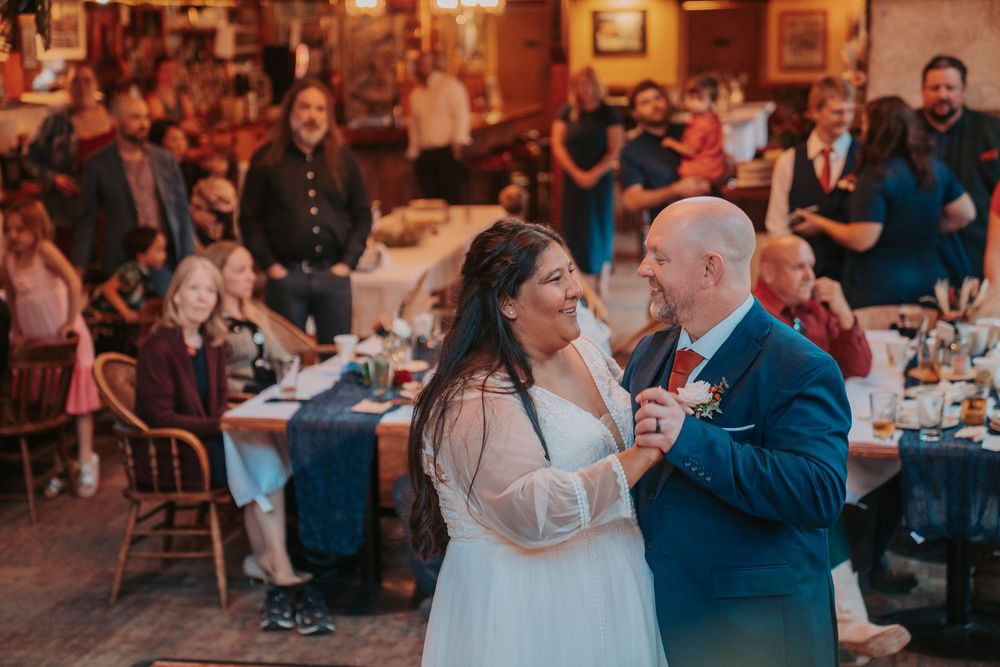 Bride and groom share first dance at rustic wedding reception with guests watching in background.