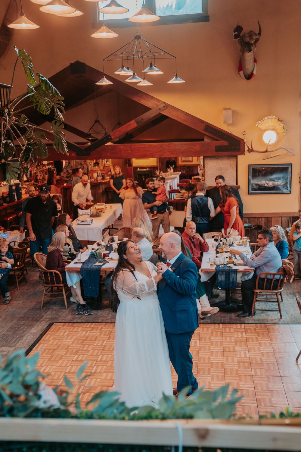 Bride and groom share first dance at rustic indoor wedding reception with guests dining at decorated tables.