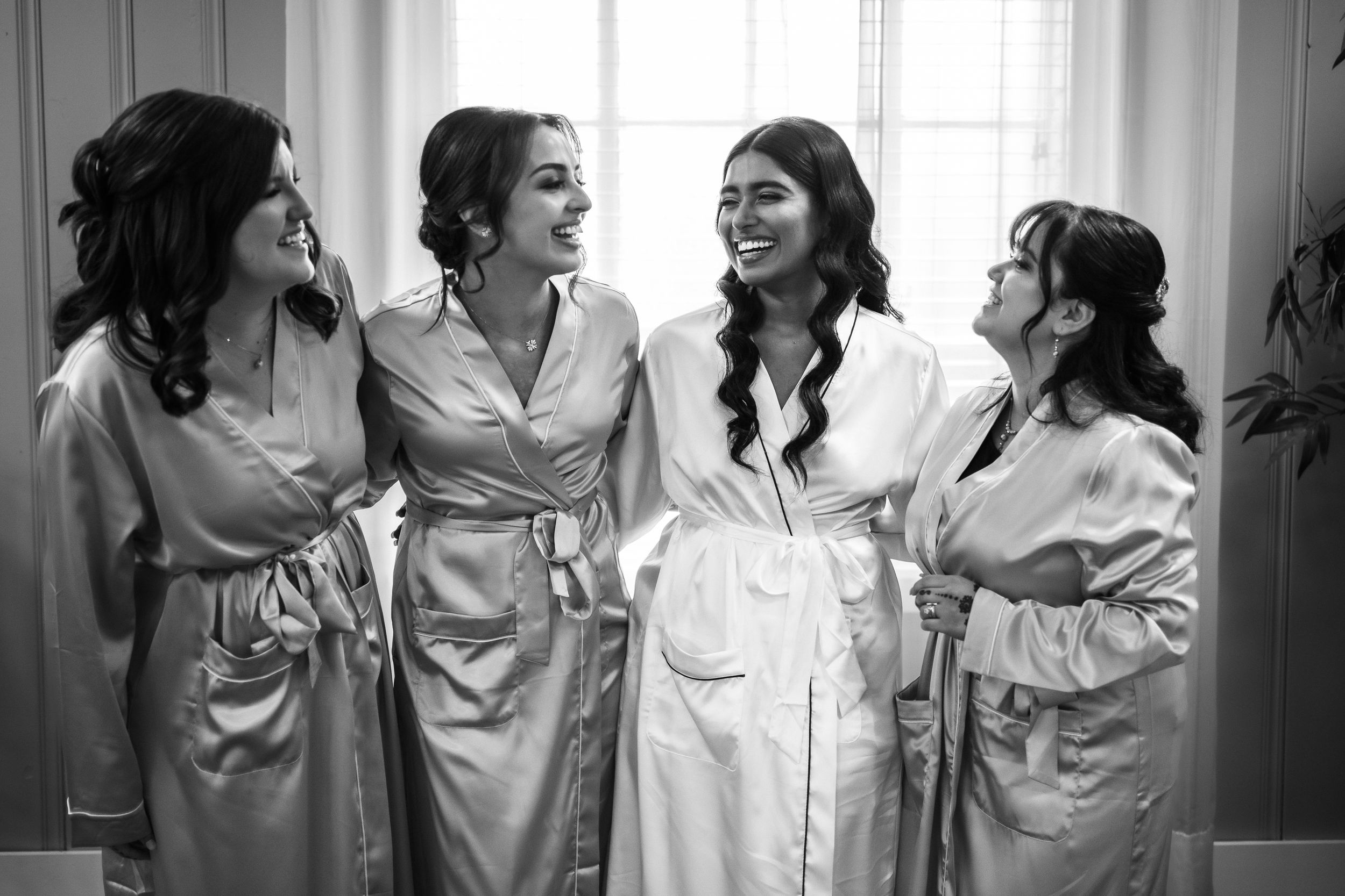 Bride and bridesmaids laughing together in satin robes during wedding morning preparations, black and white photo.