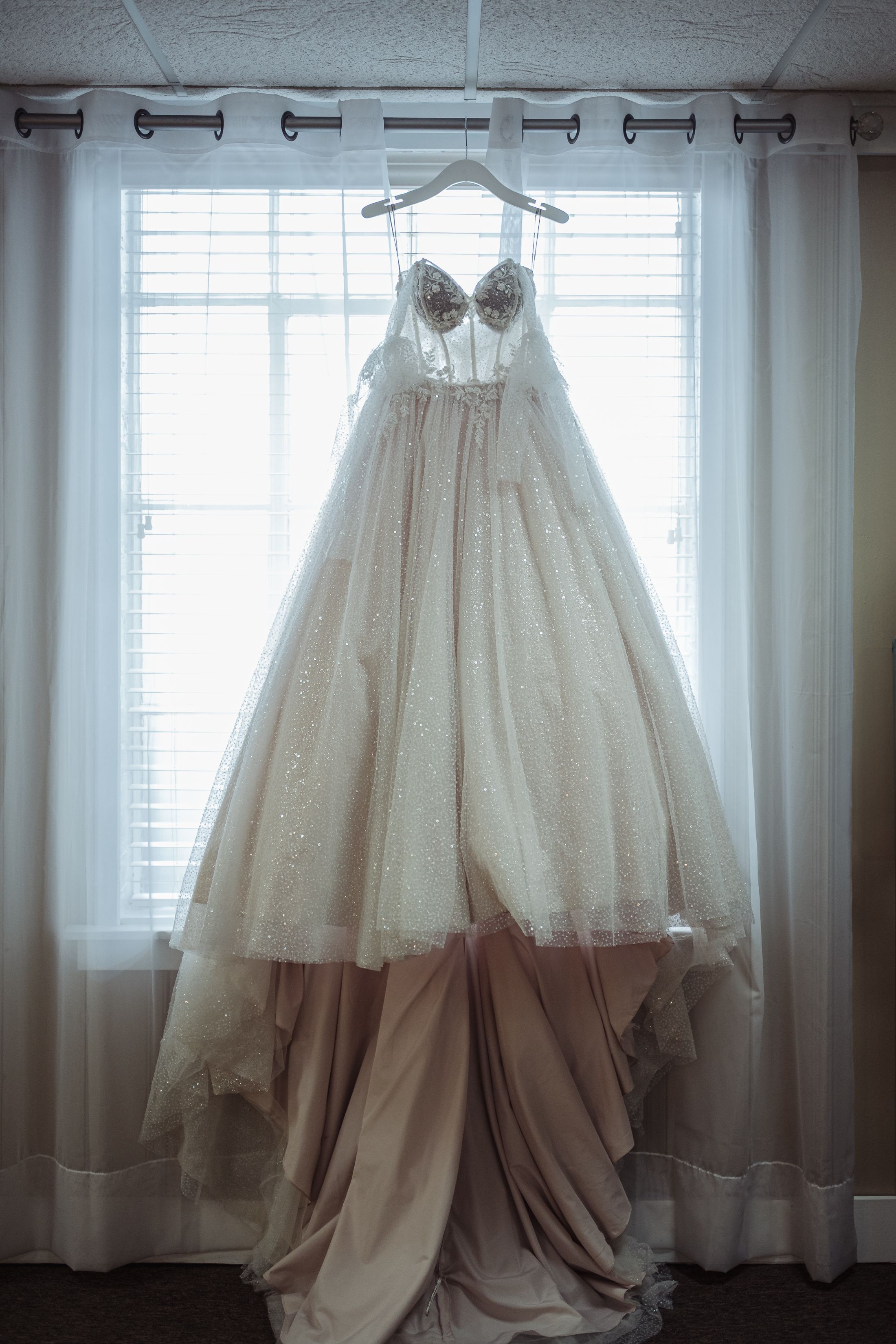 Elegant strapless ball gown wedding dress with lace bodice and champagne skirt hanging on a hanger by a bright window.