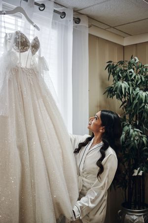 Bride in white robe admiring her sparkly ball gown wedding dress hanging by a window before her ceremony.