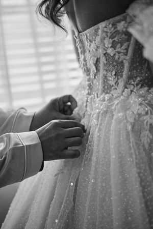 Black and white photo of hands fastening buttons on an intricate lace wedding dress with floral embroidery.