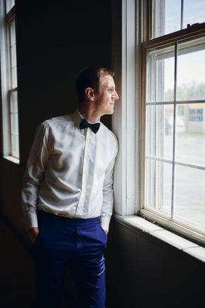Groom in white shirt and blue pants with bow tie gazes out window on wedding day.