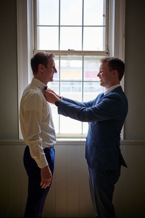 Two grooms share a joyful moment as one adjusts the other's bow tie by a sunlit window.