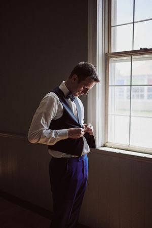 A groom in a navy vest and bow tie buttons his waistcoat by a bright window while getting ready.