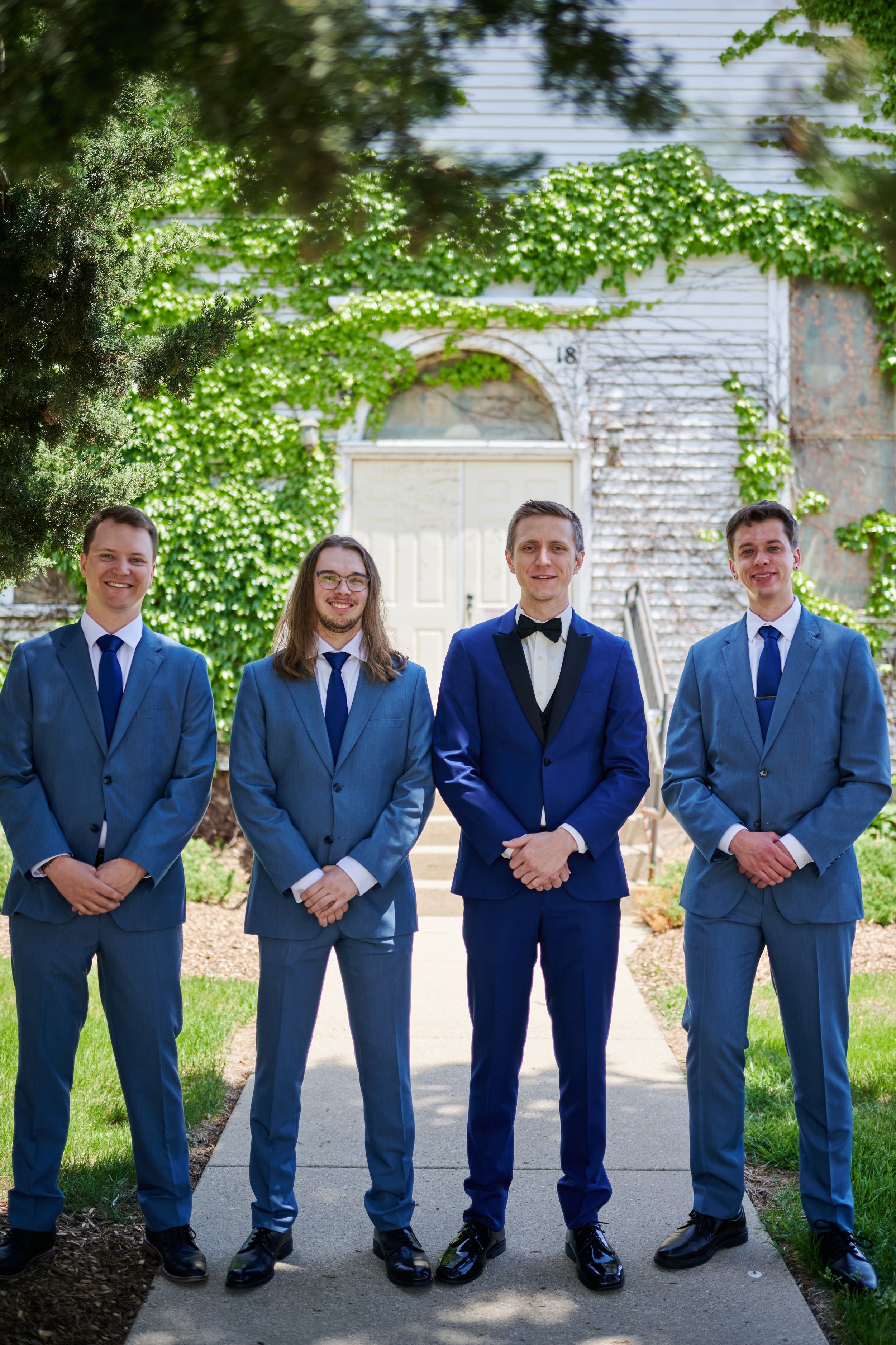 Groom in navy blue tuxedo poses with three groomsmen in light blue suits outside ivy-covered building.