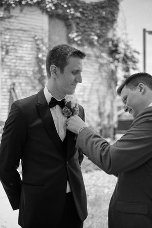Groomsman pins boutonniere on groom in black tuxedo outdoors before wedding ceremony.