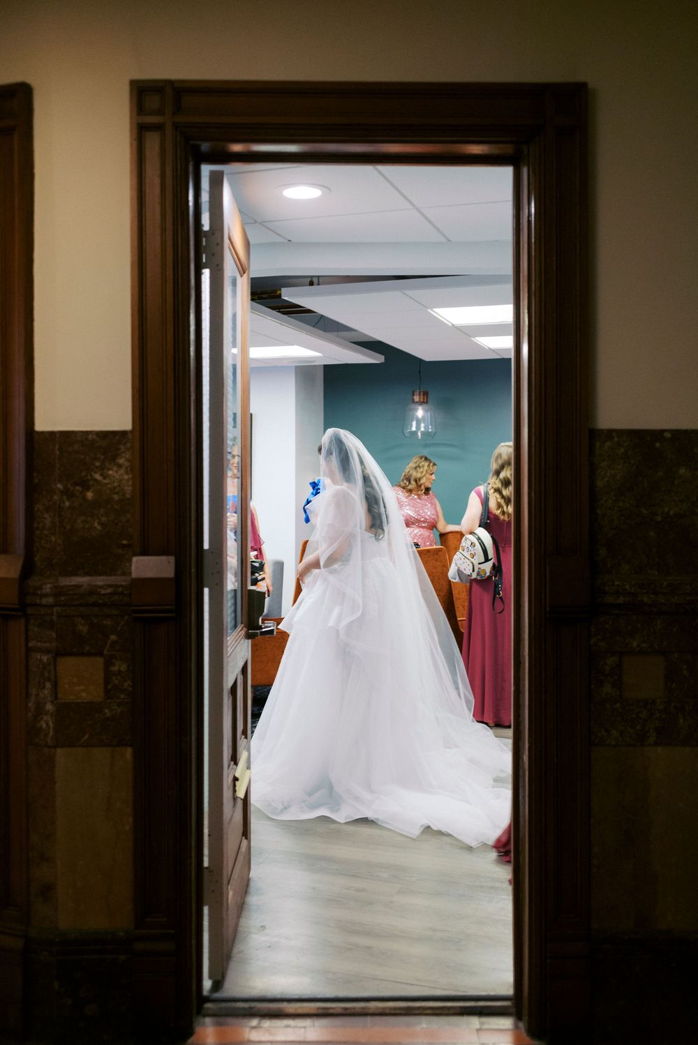 Bride in white gown and veil seen through doorway with bridesmaids in pink dresses at wedding venue.
