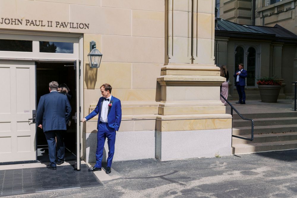 Men in formal attire outside John Paul II Pavilion, with a groom in a blue suit at a wedding venue.