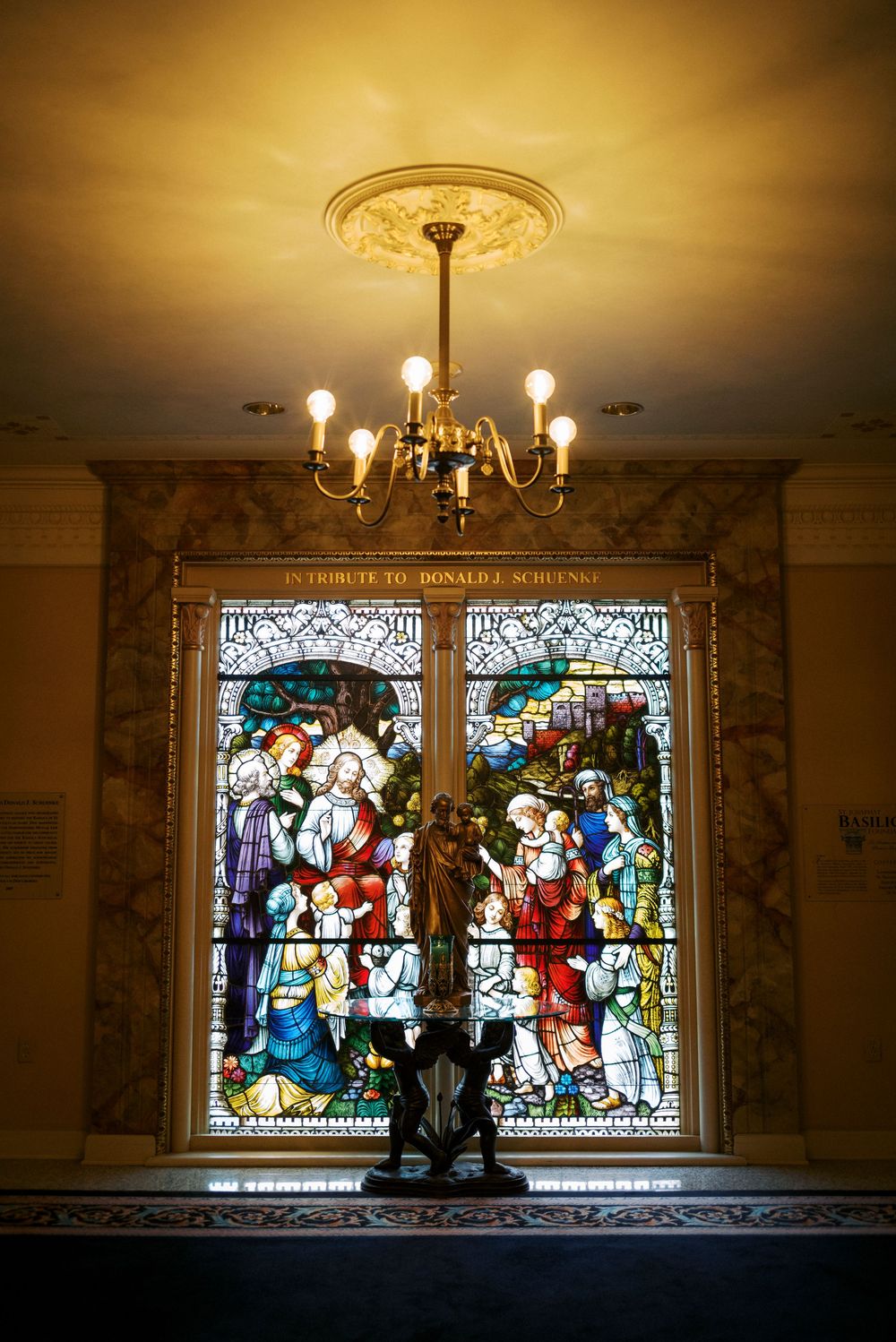 Ornate stained glass window tribute to Donald J. Scheuns illuminated by a gold chandelier in an elegant interior.