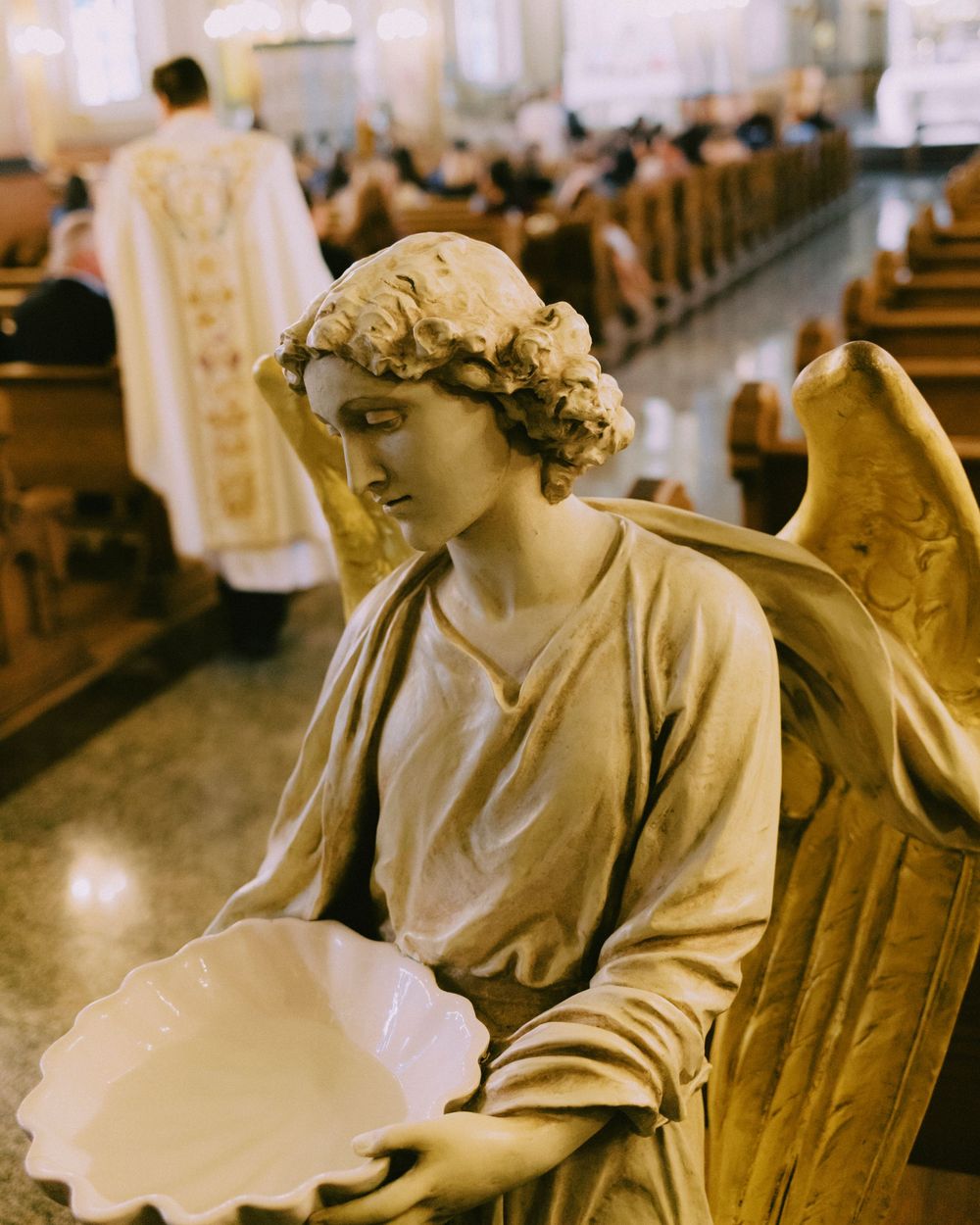Angel statue holding holy water basin inside a church during Catholic Mass with priest and congregation in background.