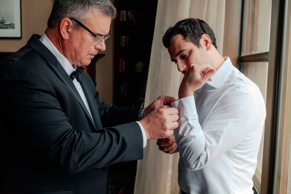 An older man in a tuxedo helps a younger man in a white dress shirt fasten his cufflinks before a wedding.