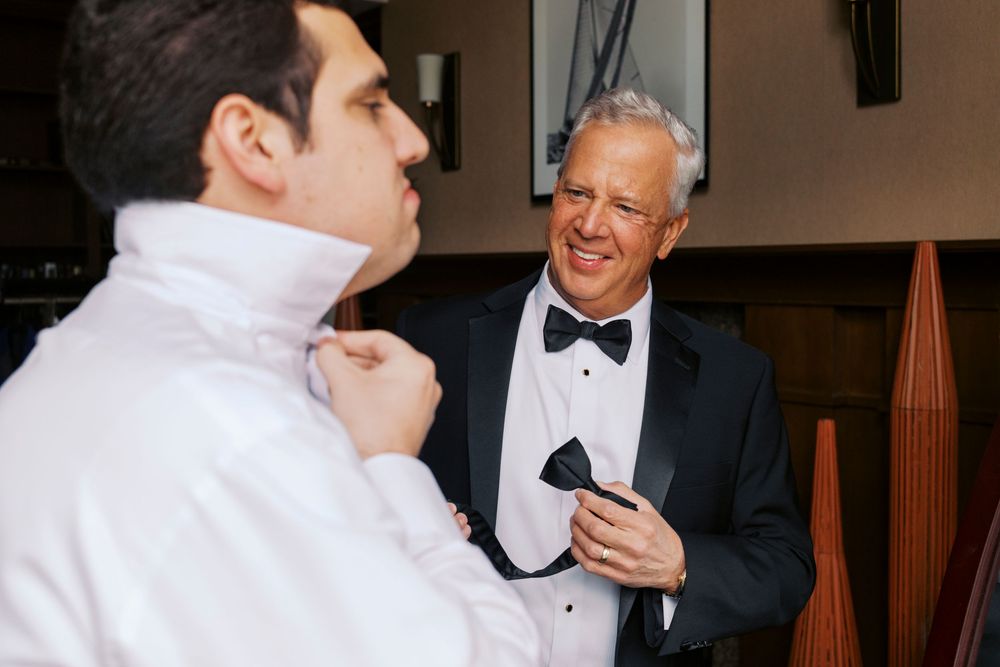 Two men in tuxedos prepare for a wedding, one adjusting his collar while the other holds a bow tie.