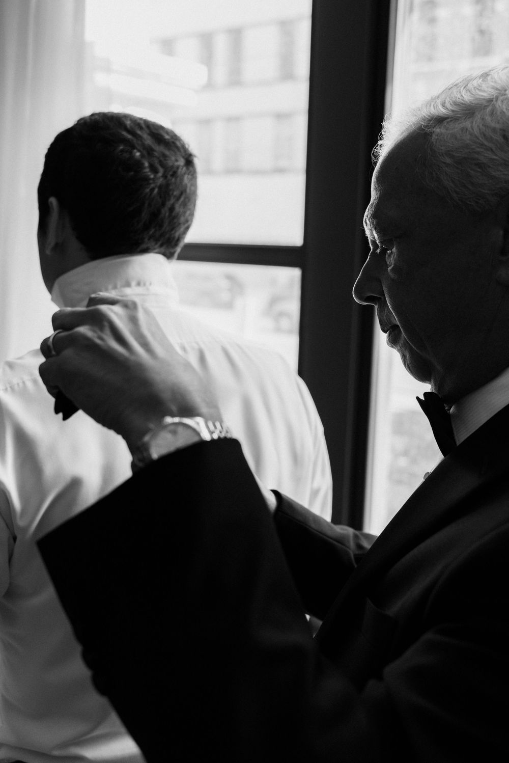 Black and white photo of an older man in a tuxedo helping a younger man button his dress shirt collar near a window.