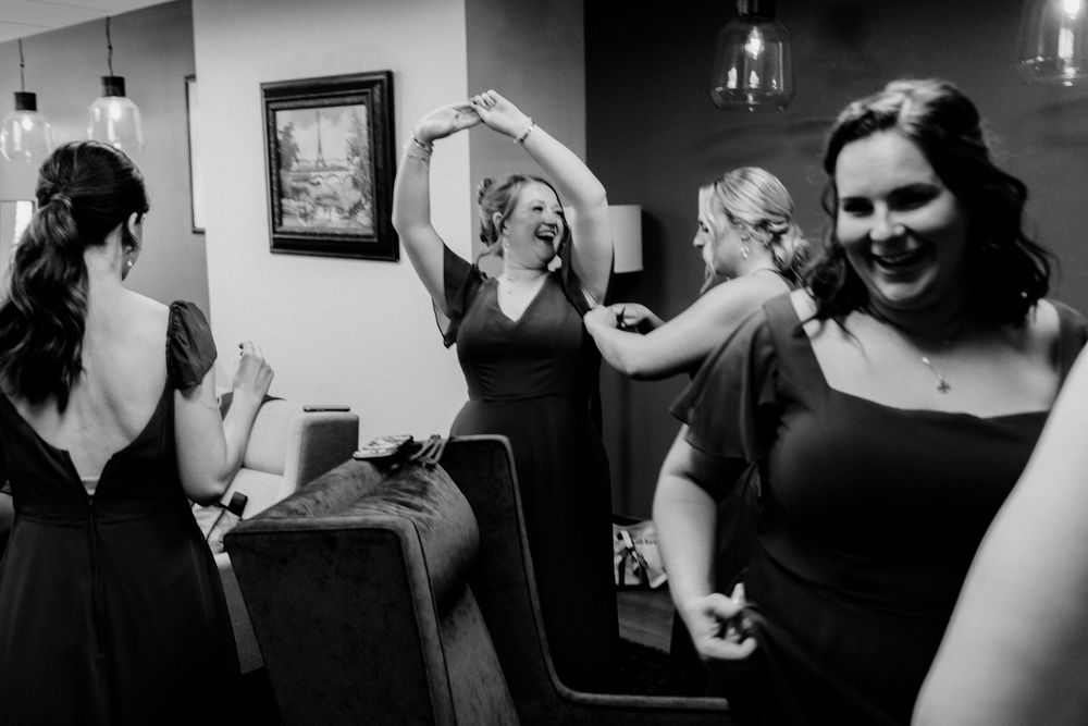 Bridesmaids dancing and laughing joyfully in black dresses during wedding celebration in black and white photo.