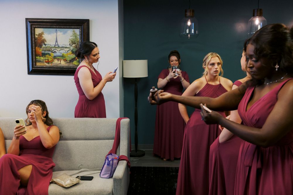 Bridesmaids in burgundy dresses check phones and touch up makeup in a teal-walled bridal suite before the wedding.