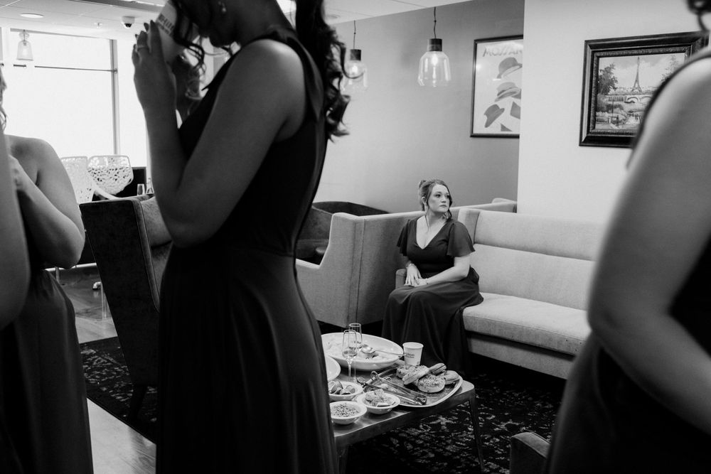Black and white photo of bridesmaids in dark dresses preparing, with a woman relaxing on a sofa nearby.