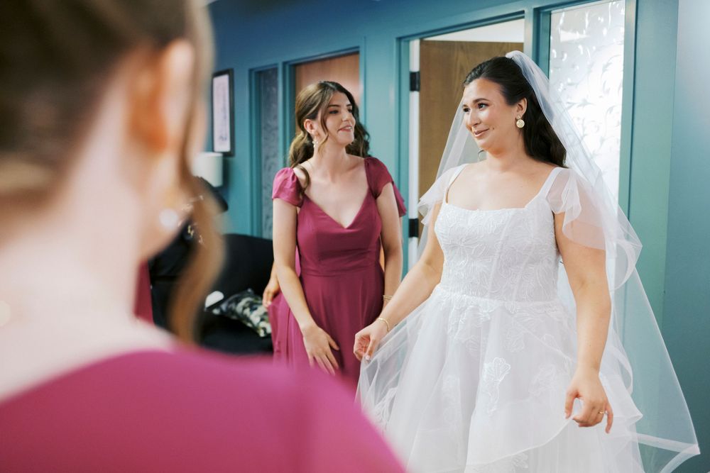 Bride in white gown and veil smiles at bridesmaid in burgundy dress during wedding preparations.
