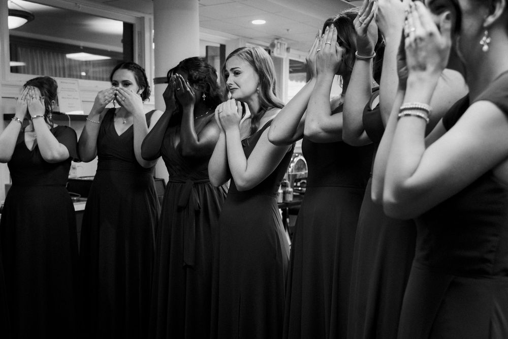 Emotional bridesmaids in dresses covering their faces while crying during a dress reveal, black and white candid photo.