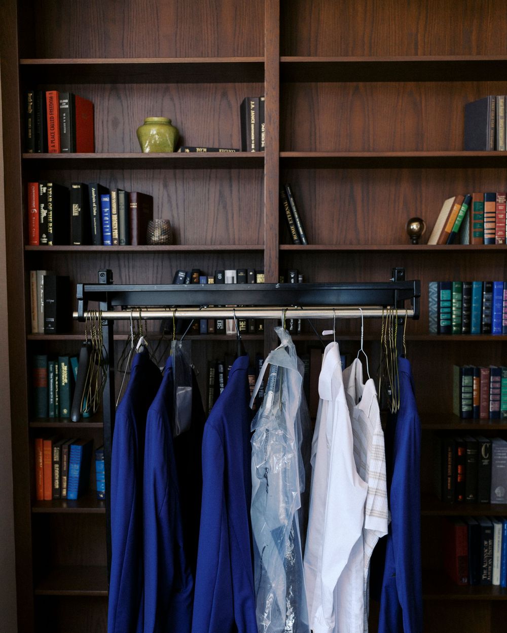 Clothing rack with blue and white shirts in front of dark wood bookshelves filled with books and decor.