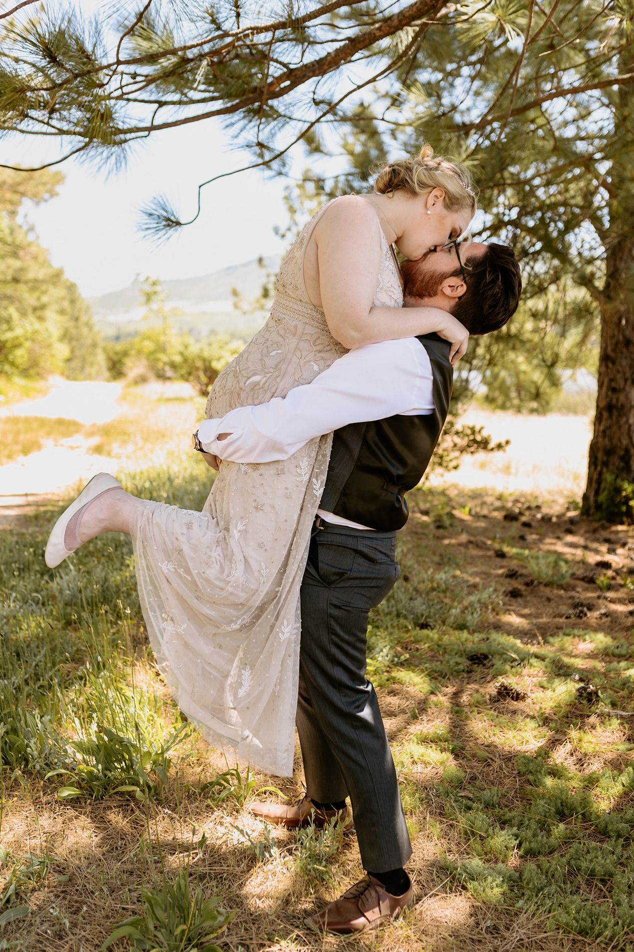 Groom lifting bride for romantic kiss outdoors under pine trees on sunny wedding day at Lake Cuyamaca, Julian.