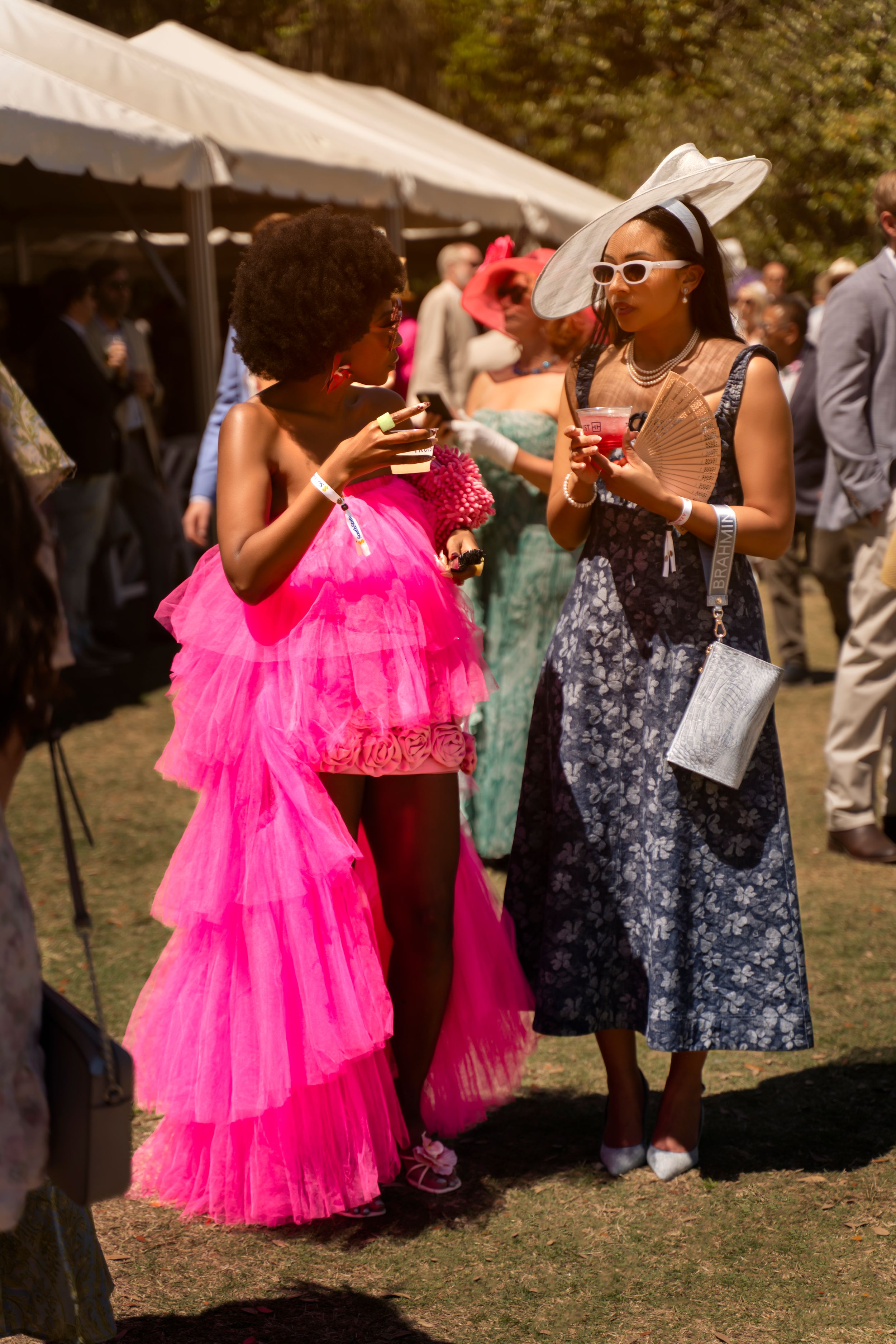 Two women in colorful outfits, one in a pink tulle gown, socializing at an outdoor garden party event.