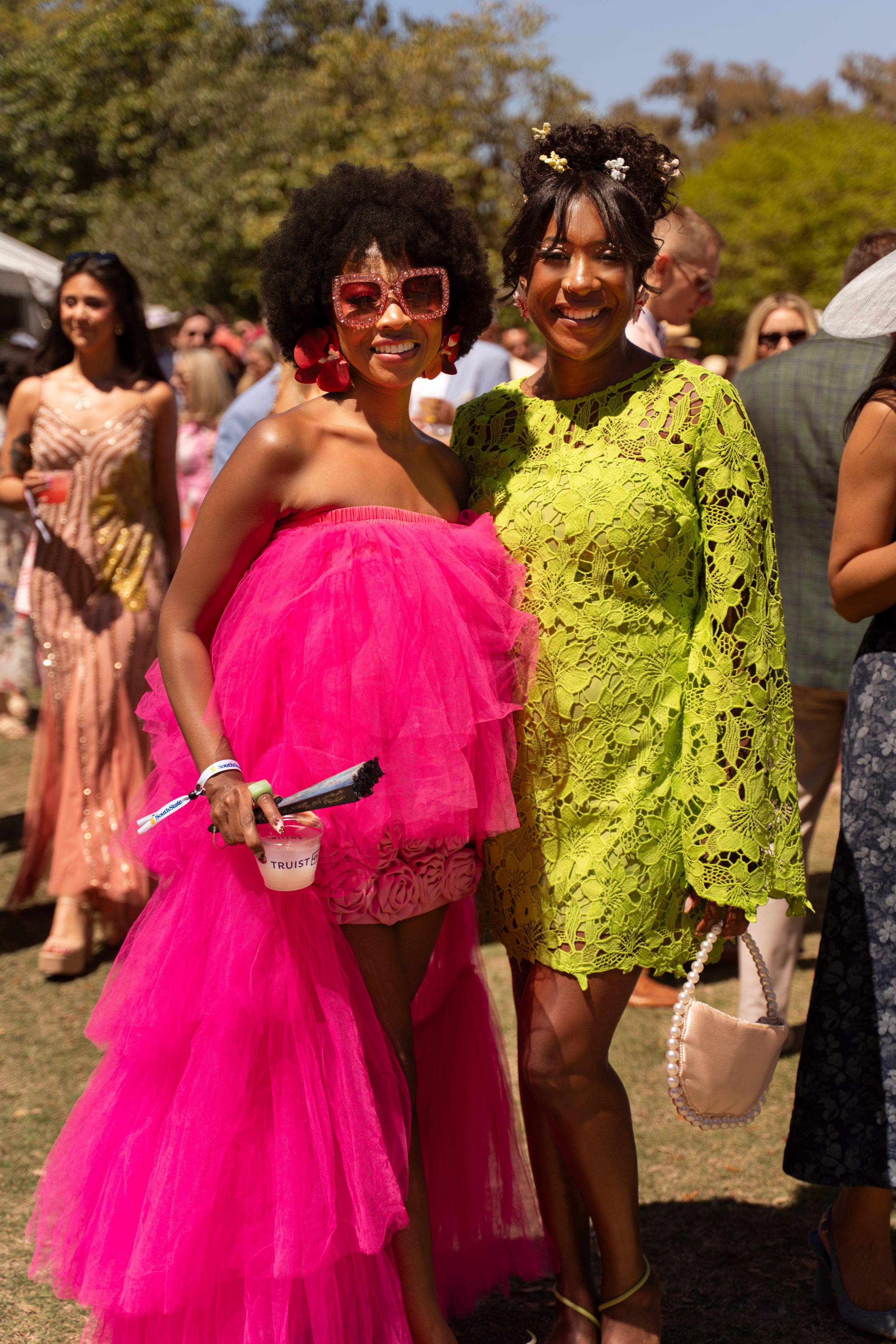 Two women pose outdoors, one in a pink tulle gown and one in a yellow lace dress, at Airlie Gardens, Wilmington NC, Azalea Fest Garden Party