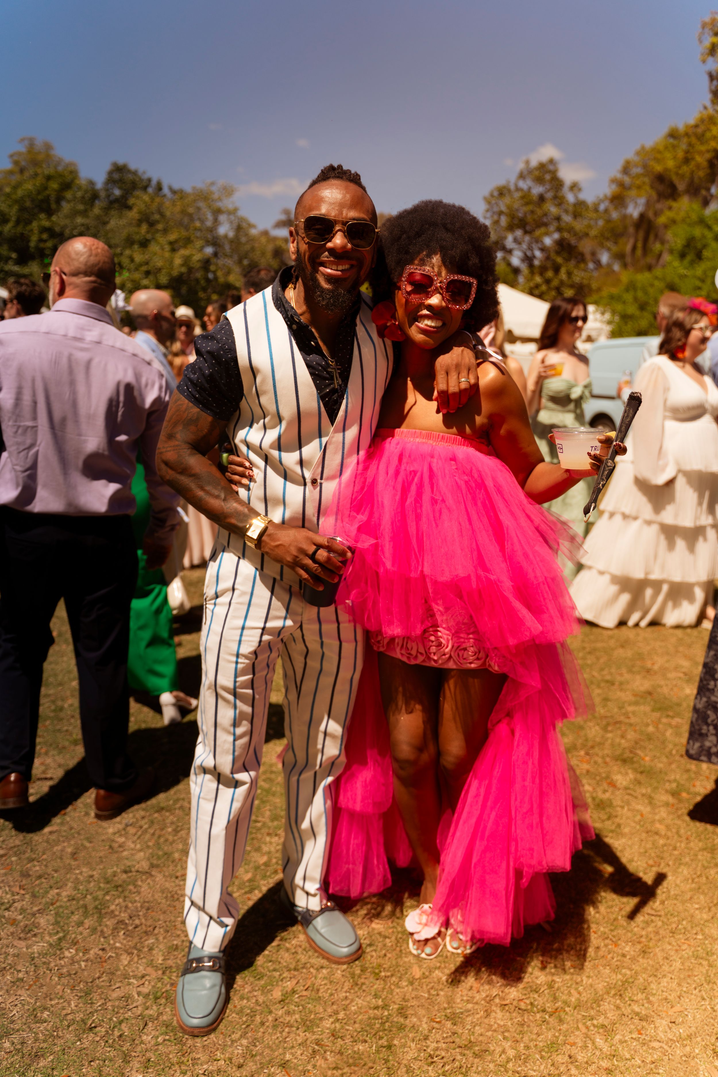 Stylish couple posing at outdoor garden party, man in striped suit, woman in vibrant pink tulle dress with afro and sunglasses.
