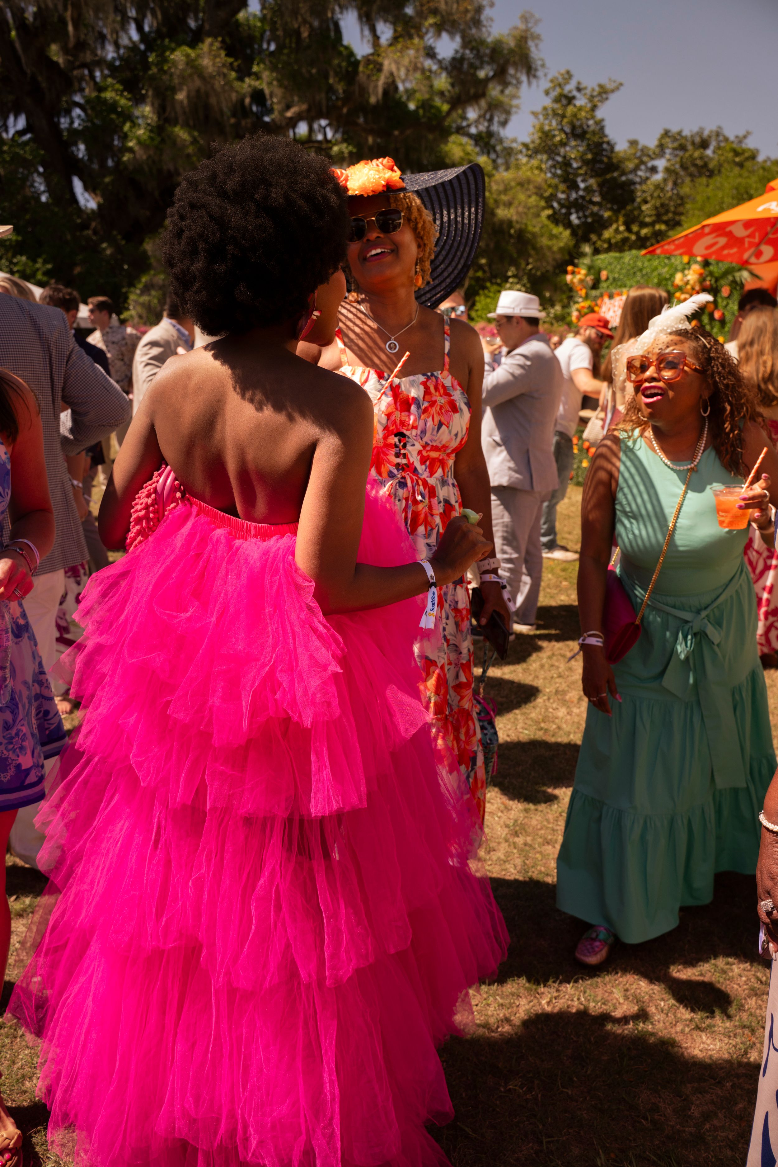 Women laughing and socializing at elegant outdoor event, one wearing a striking hot pink ruffled tulle gown.