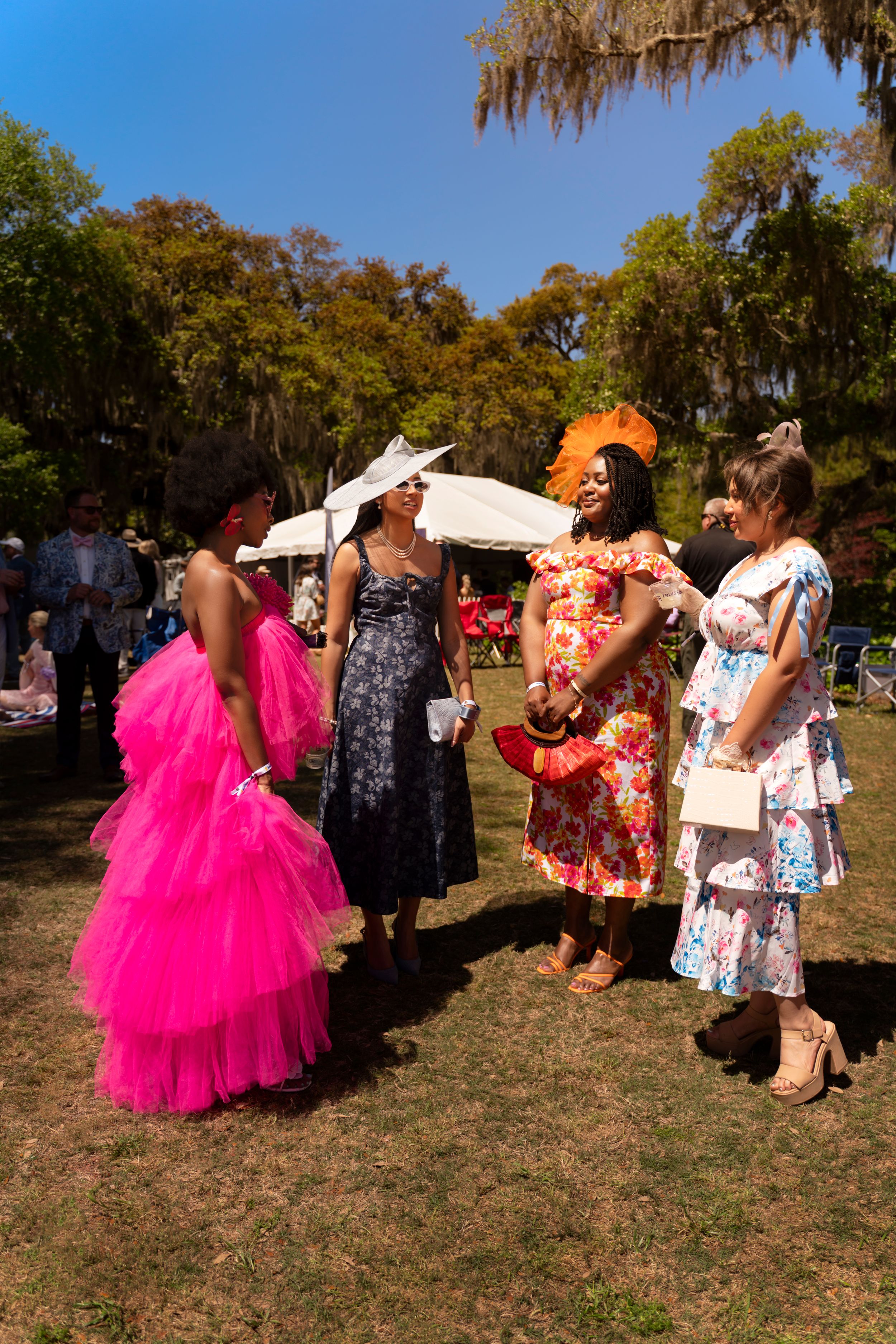 Four elegantly dressed women in colorful gowns and fascinators socializing outdoors at a garden party event.