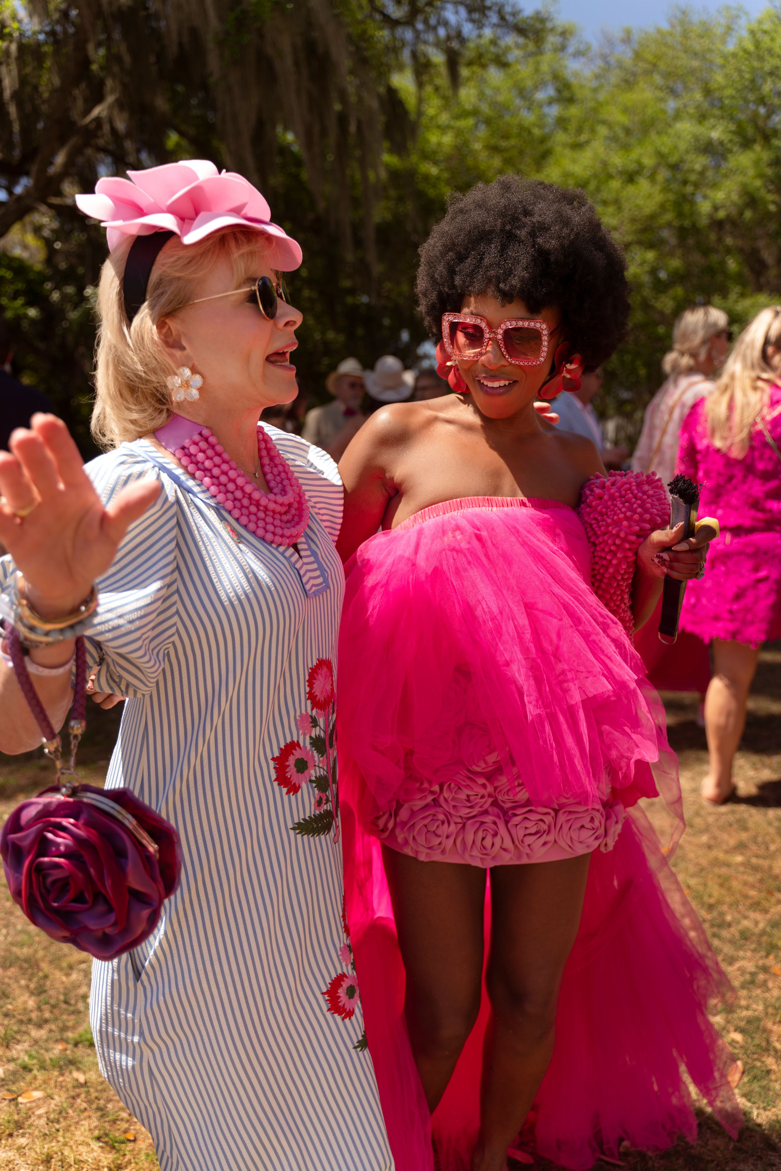Two women in vibrant pink outfits and accessories smile together at an outdoor garden party event.