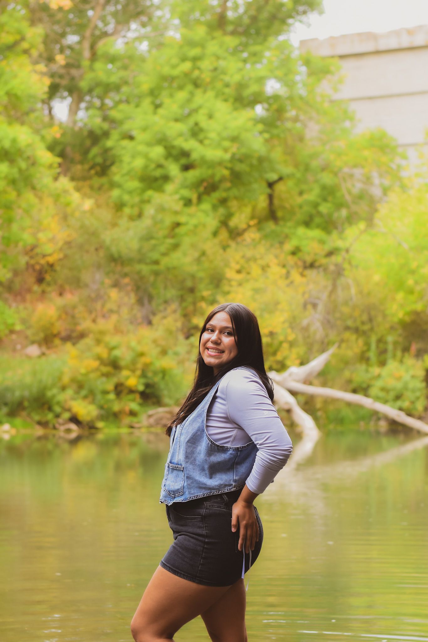 Young woman in denim vest and black skirt smiling by a serene pond surrounded by lush green autumn trees.
