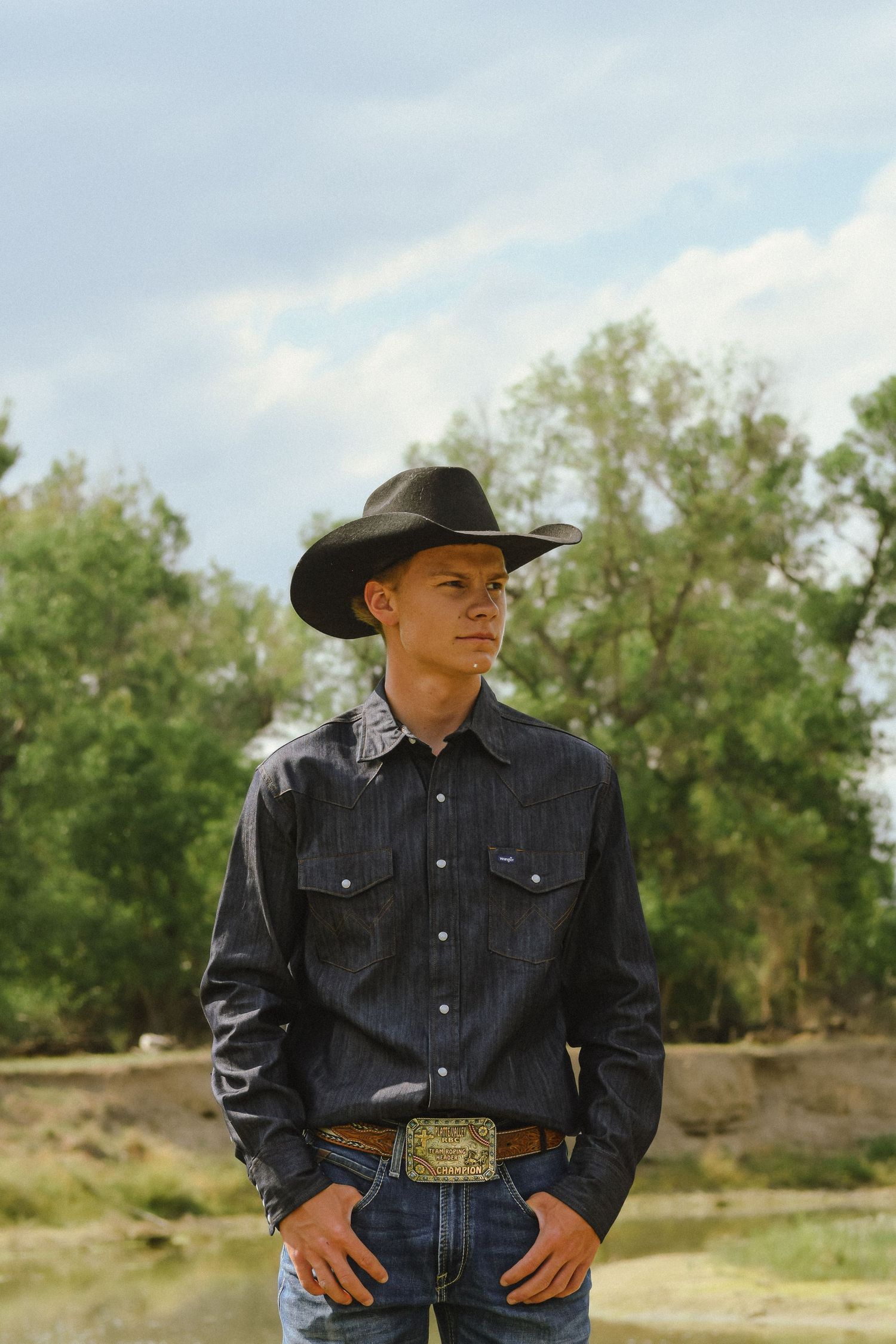 Woman in black cowboy hat holding cameras outdoors near bare trees in a rural setting.