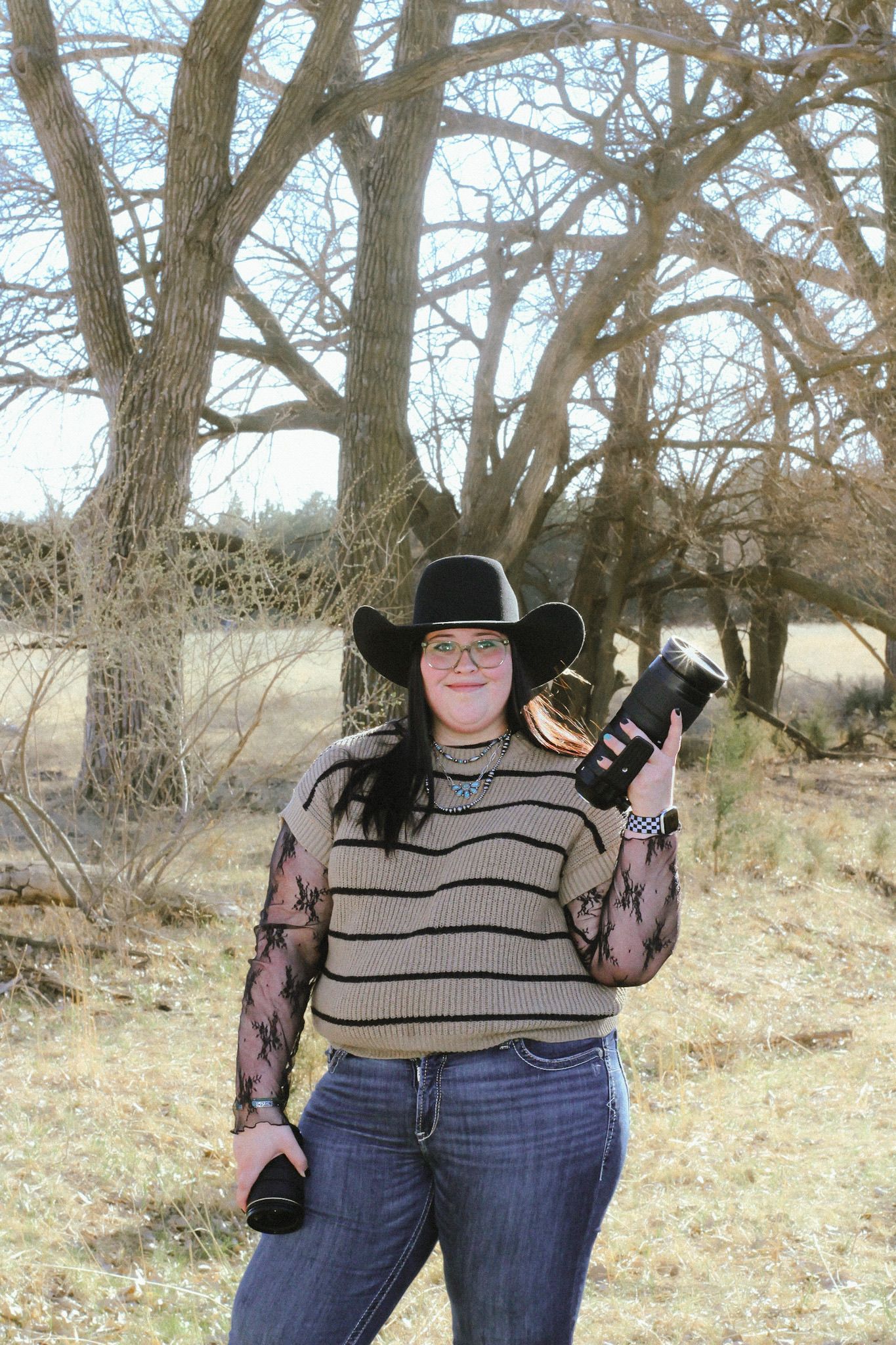 Woman in black cowboy hat holding cameras outdoors near bare trees in a rural setting.
