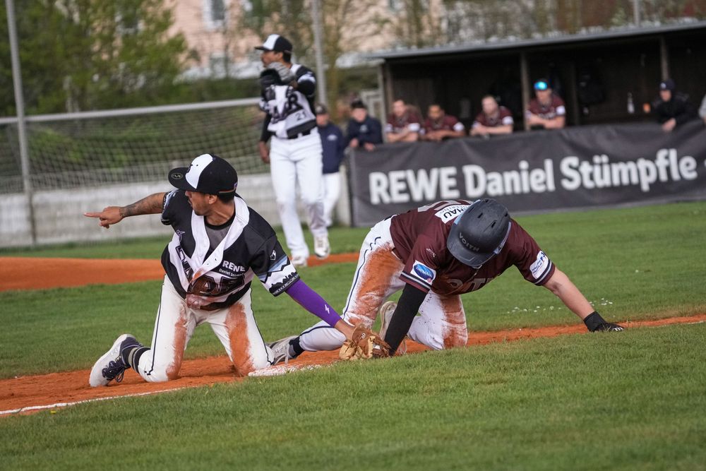 Baseball player slides into base as fielder attempts tag during a competitive game near REWE Daniel Stümpfle signage.