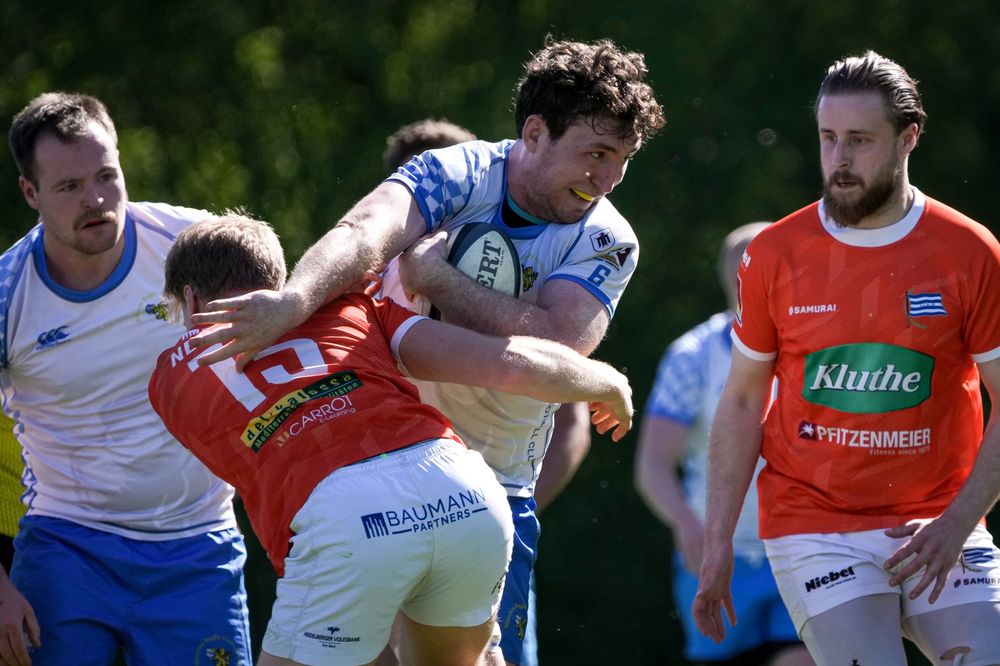 Rugby players in blue and orange jerseys compete in an intense tackle during an outdoor match on a green field.