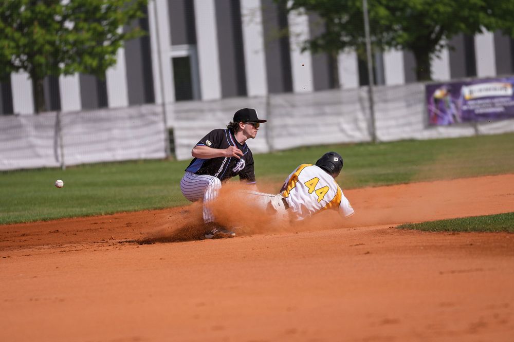 Baseball player sliding into base as infielder catches ball during competitive game on dirt infield.