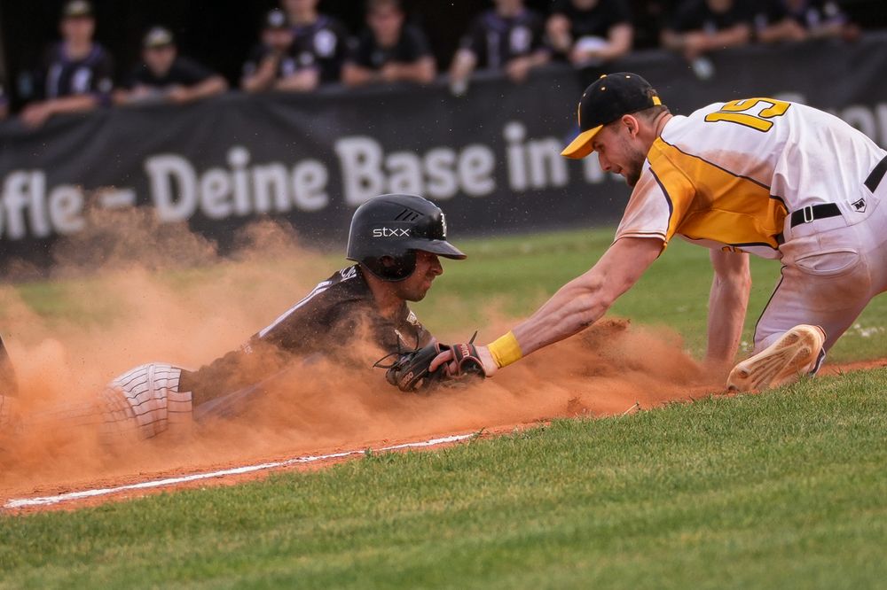 Baseball player slides into base as fielder attempts tag play in a cloud of orange dirt during a night game.