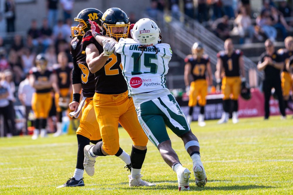 Two American football players compete during a sunny outdoor game, one in black and gold, the other in green and white jersey number 15.