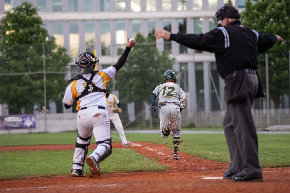 Baseball catcher awaits throw at home plate as runner #12 approaches, with umpire signaling during a competitive game.