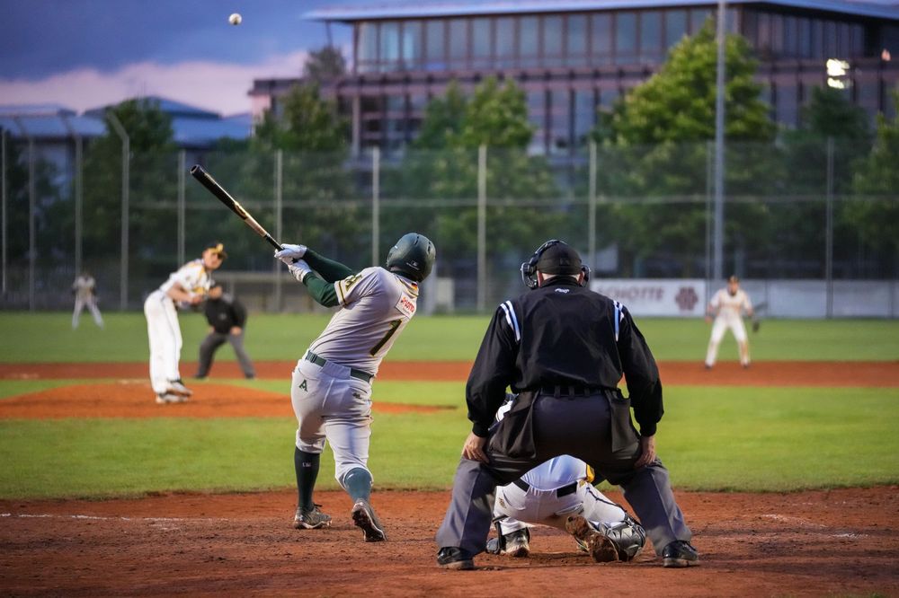 Baseball batter swinging at pitch with catcher and umpire crouching behind home plate on an outdoor dirt field.