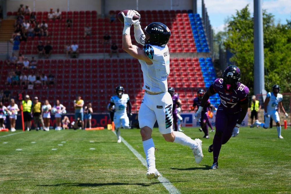 American football player catches a pass near the end zone during a sunny outdoor game, defender in purple trails behind.