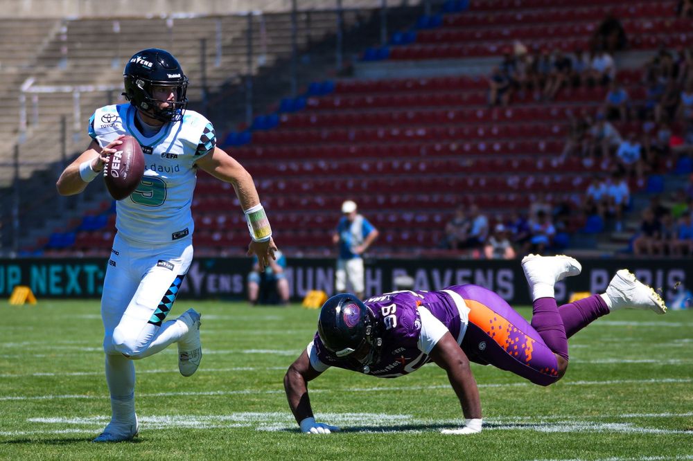 American football player in teal uniform evades a diving defender in purple during a sunny outdoor game.