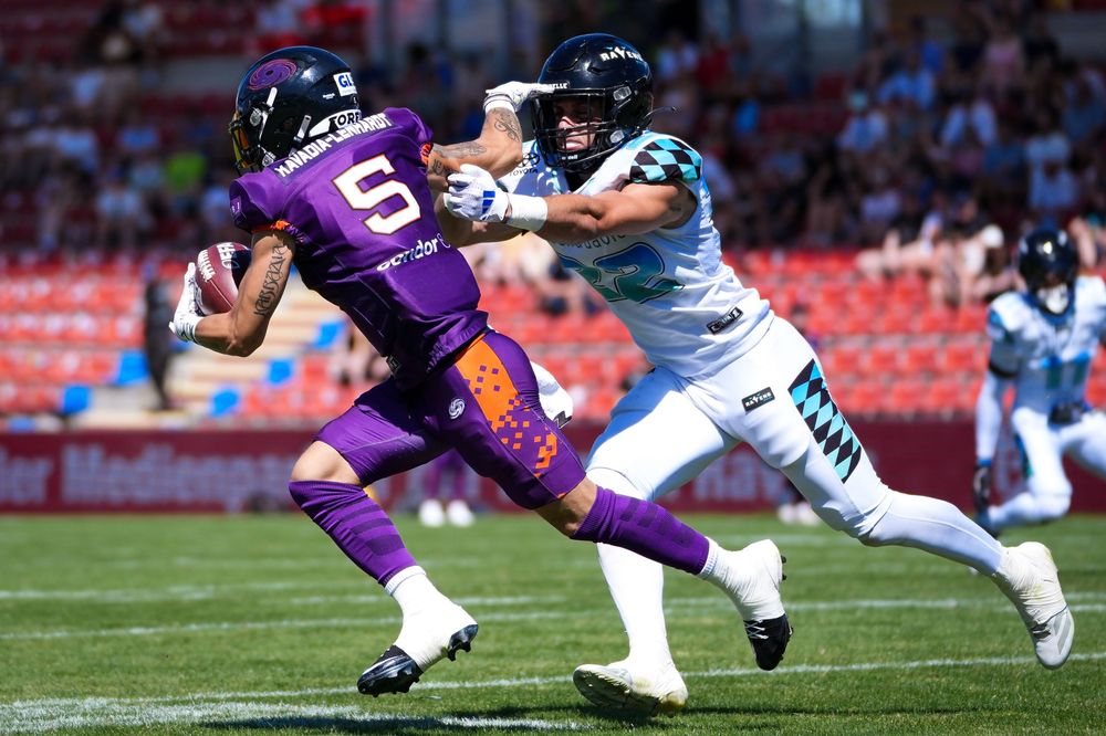 Football player in purple jersey number 5 evades a tackle during an outdoor arena football game.