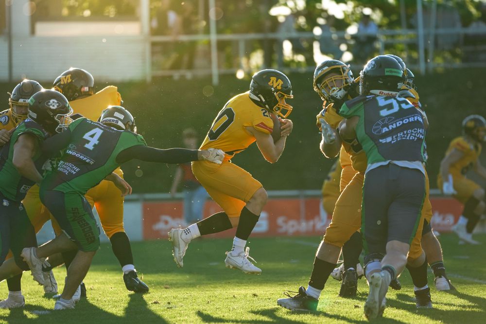 American football players in yellow and green uniforms compete in an intense game under bright evening sunlight.