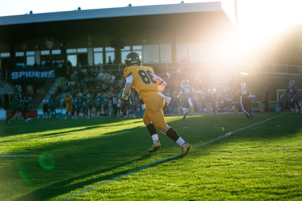 Football player wearing #86 yellow jersey runs on field at sunset with packed Spiders stadium stands in background.