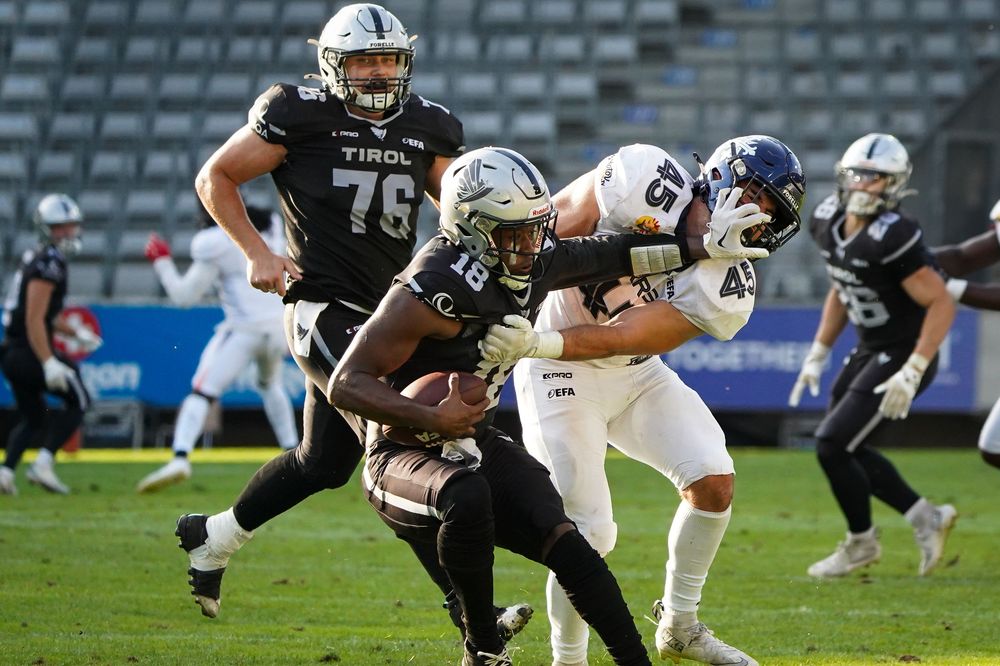 American football player carrying ball evading tackle during Tirol Raiders game on green field.
