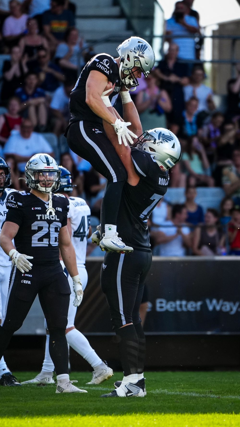 Two American football players in black Tirol uniforms celebrate a touchdown, with one lifting the other in the air.